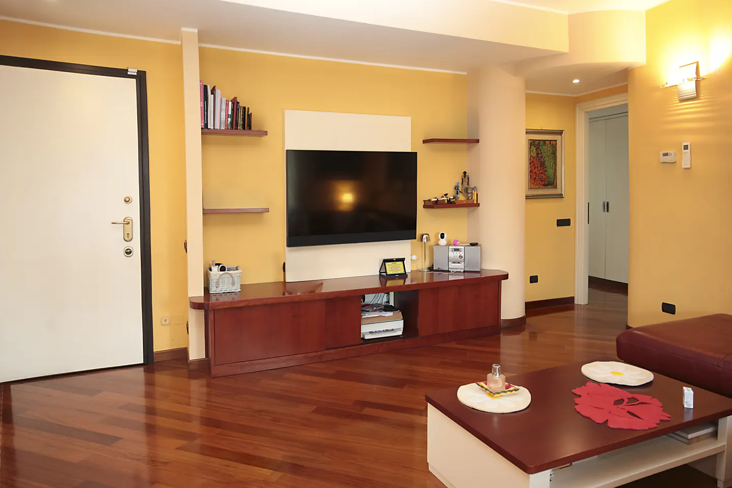Living room with yellow walls, wood floors, and a large TV. Shelves hold books and decor. A coffee table sits in front of a red leather couch.
