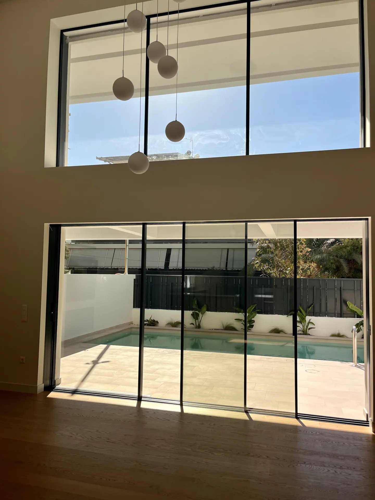 Interior view of a modern home with large windows overlooking a pool and backyard. White pendant lights hang from the ceiling.