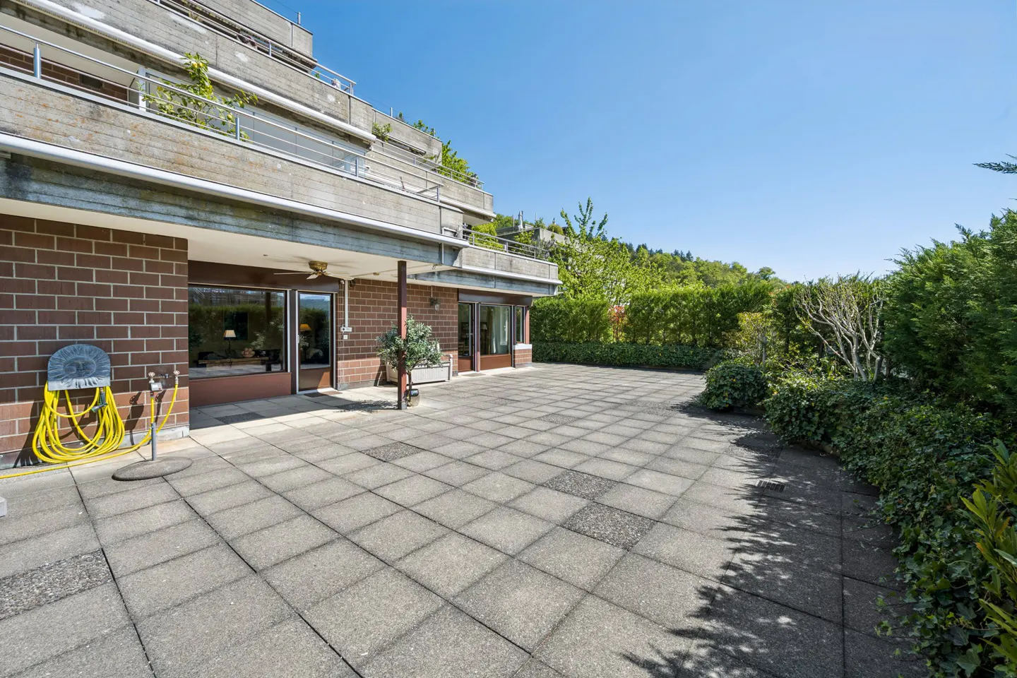 Wide patio with gray pavers outside a brick and concrete building. A yellow hose is coiled on the left. Green hedges line the patio's edge.