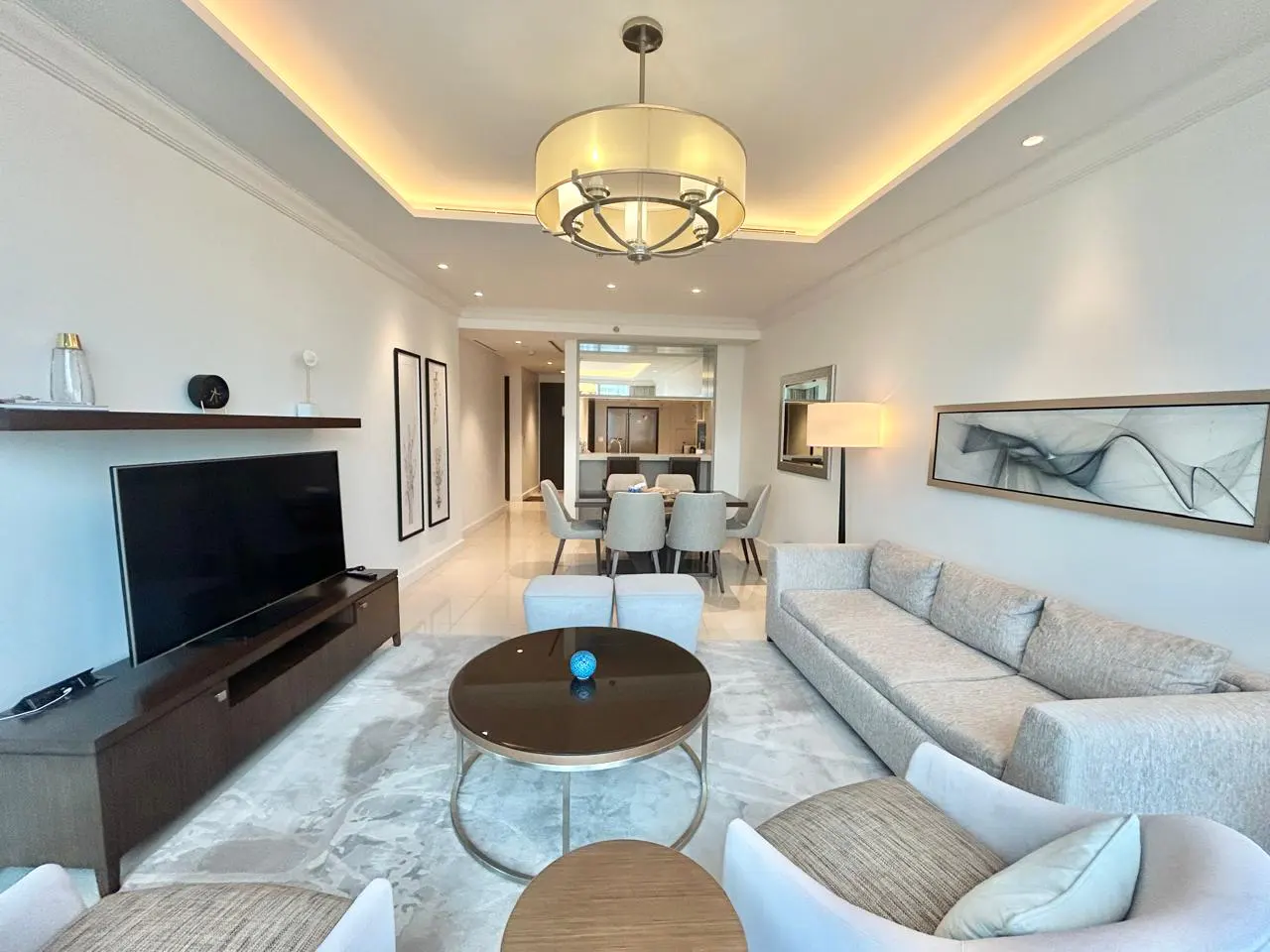 A bright, modern living room with a gray sofa, white chairs, and a round glass coffee table. A dining area and kitchen are visible in the background.