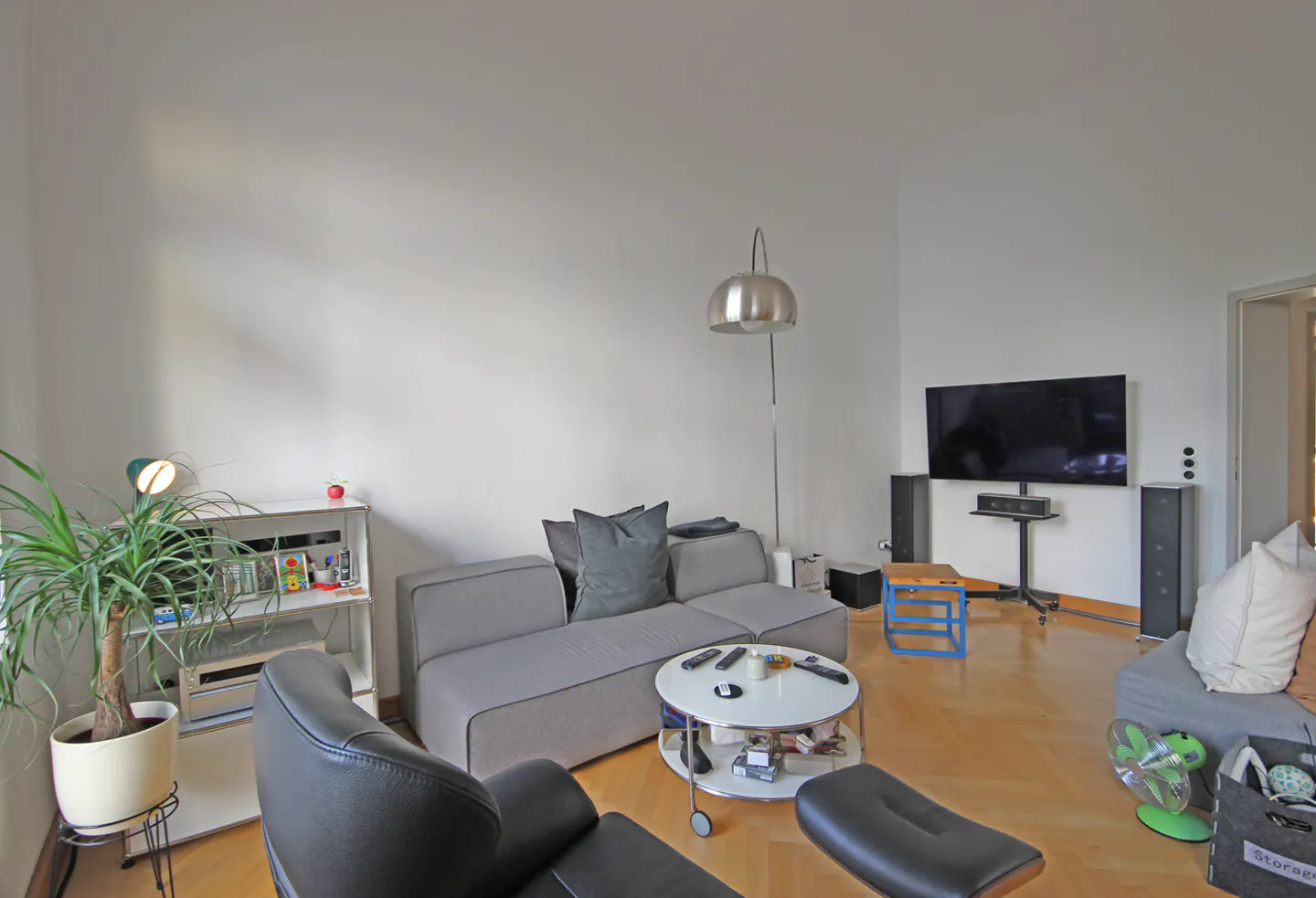 Living room with gray sofa, black chair, and TV. A floor lamp stands near the sofa. A potted plant sits on a shelf.
