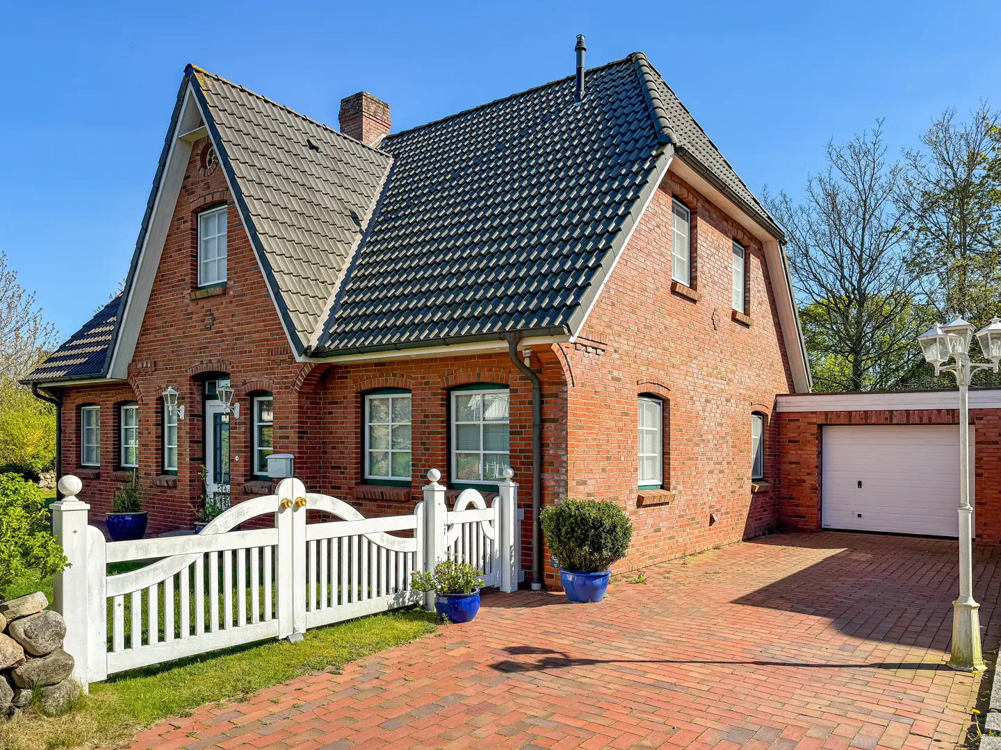 A two-story red brick house with a gray tiled roof, white fence, and brick driveway on a sunny day.