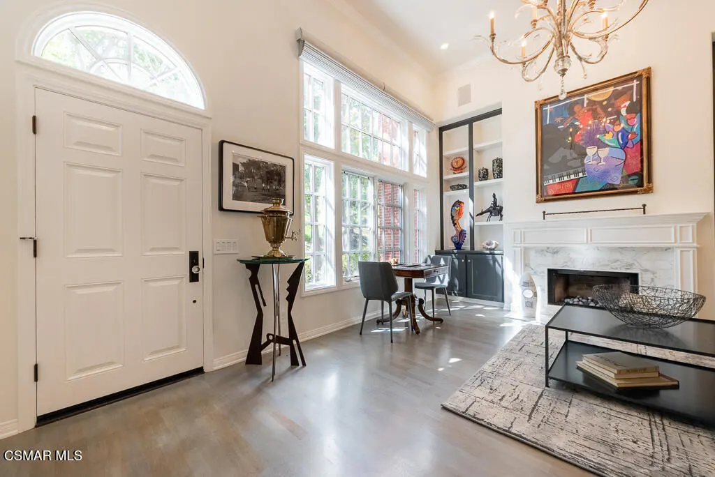 Bright living room with white walls, a fireplace, and large windows. A white door is on the left, and a chandelier hangs from the ceiling.
