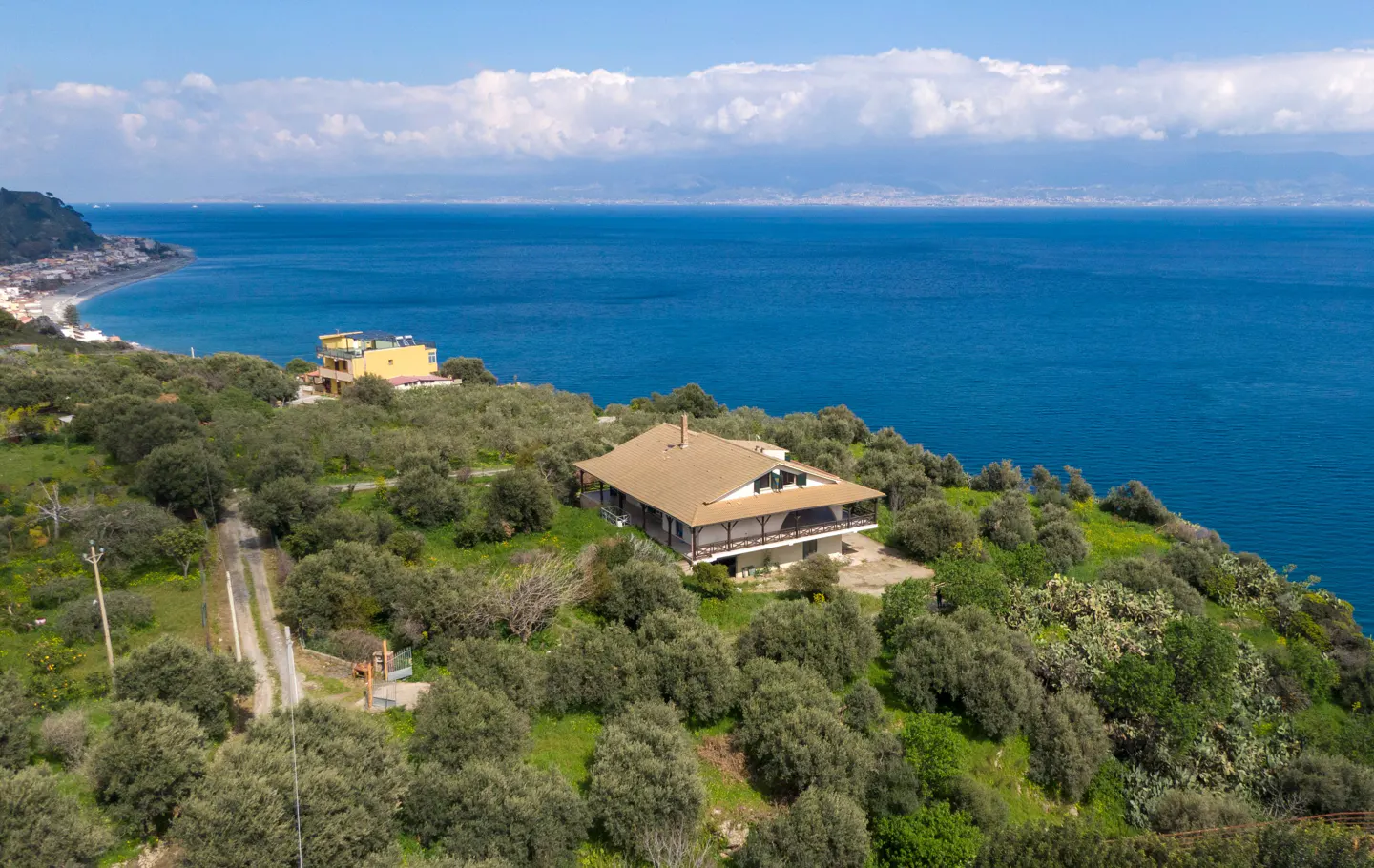 Aerial view of a tan house with a porch on a green hill overlooking the blue ocean and a town on the coast.