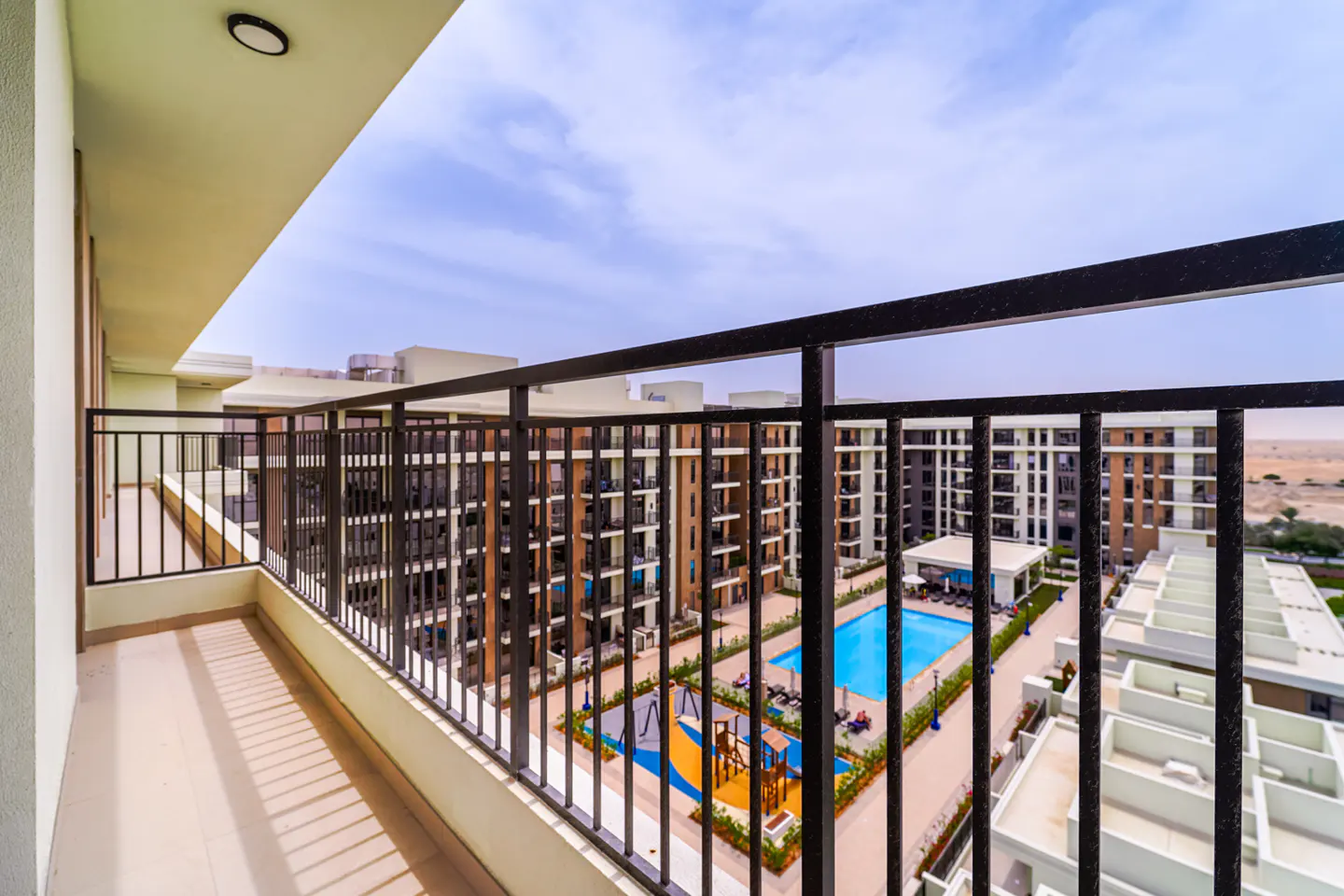 View from a balcony with black railings overlooking a pool, playground, and apartment buildings under a blue sky.