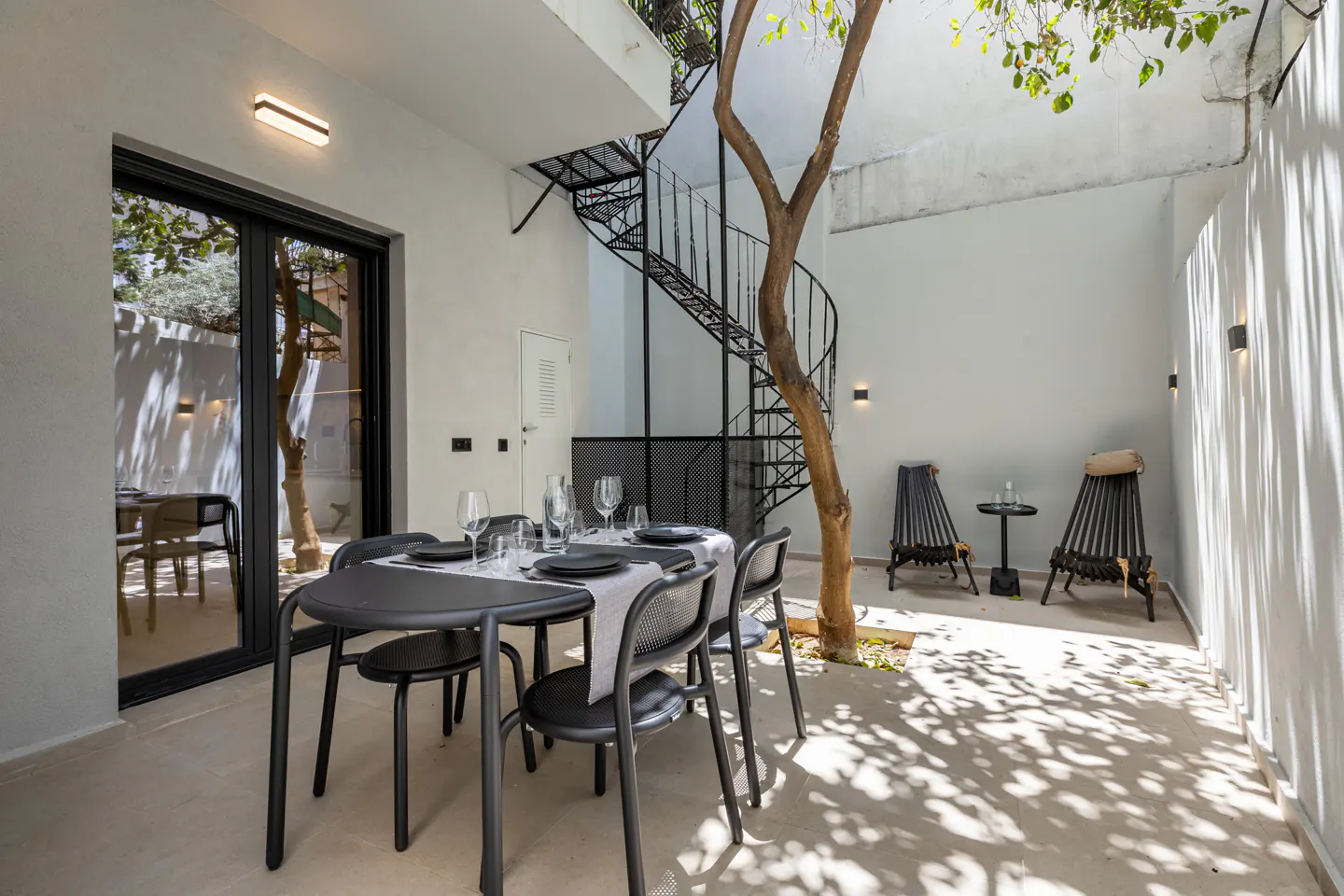 Outdoor dining area with a black table set for six, chairs, and a spiral staircase. A tree grows in the center, casting shadows on the white walls and floor.