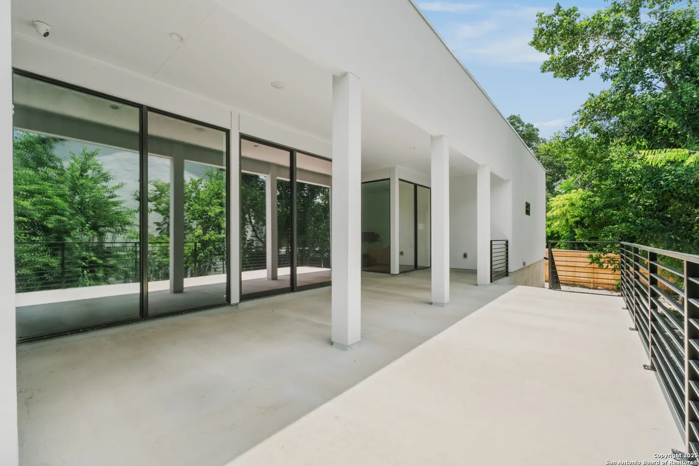 Exterior view of a modern white house with large windows and columns, a concrete patio, and a black metal railing. Green trees are visible in the background.