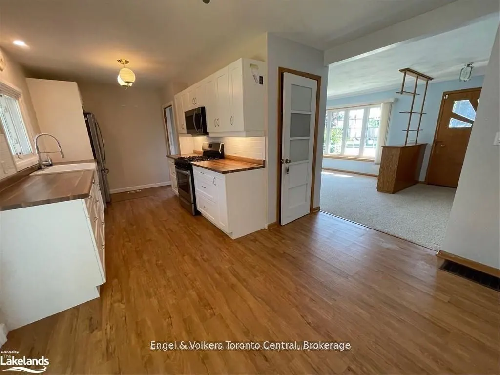 A kitchen with wood floors, white cabinets, and butcher block countertops. A doorway leads to a living room with a front door.