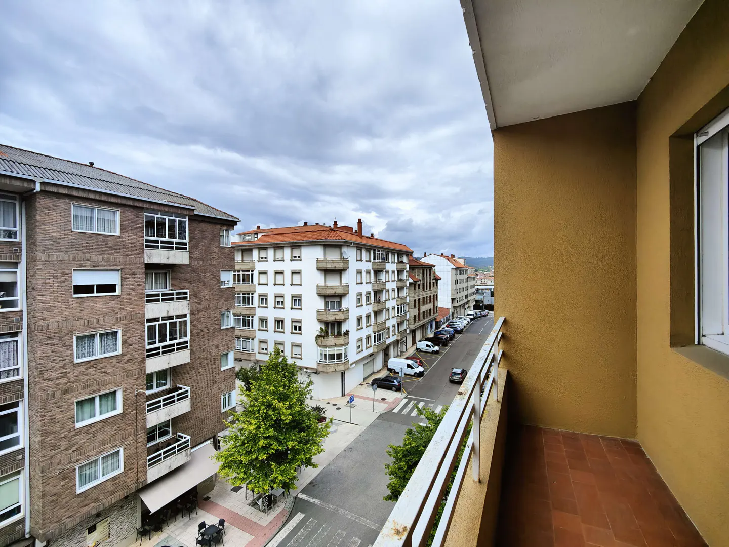 View from a balcony with white railings overlooking a city street with parked cars and apartment buildings under a cloudy sky.
