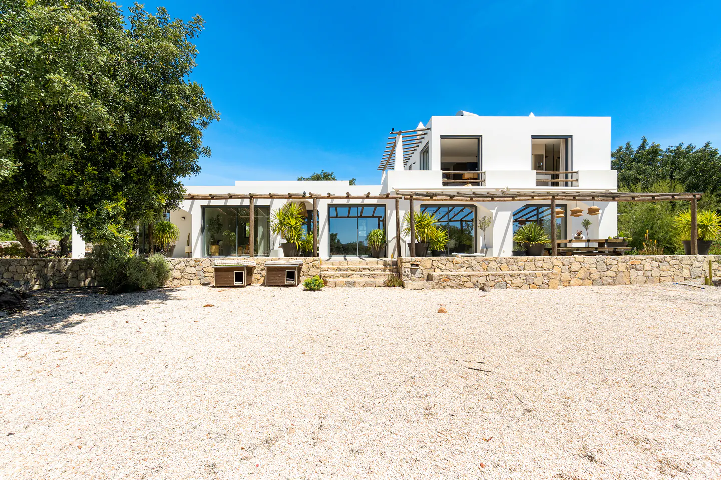 Modern white two-story villa with a stone base, gravel yard, and wooden pergola under a clear blue sky.