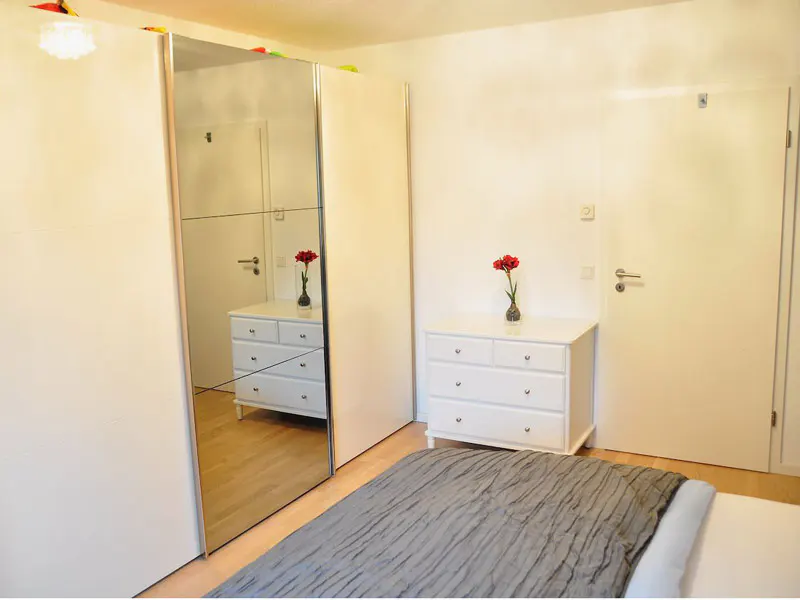 Bedroom with a gray bedspread, white dresser with red flowers, and a mirrored closet.