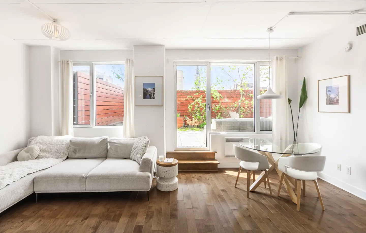 Bright living room with a white couch, glass table, and wood floors. A door leads to a patio with a red brick wall.