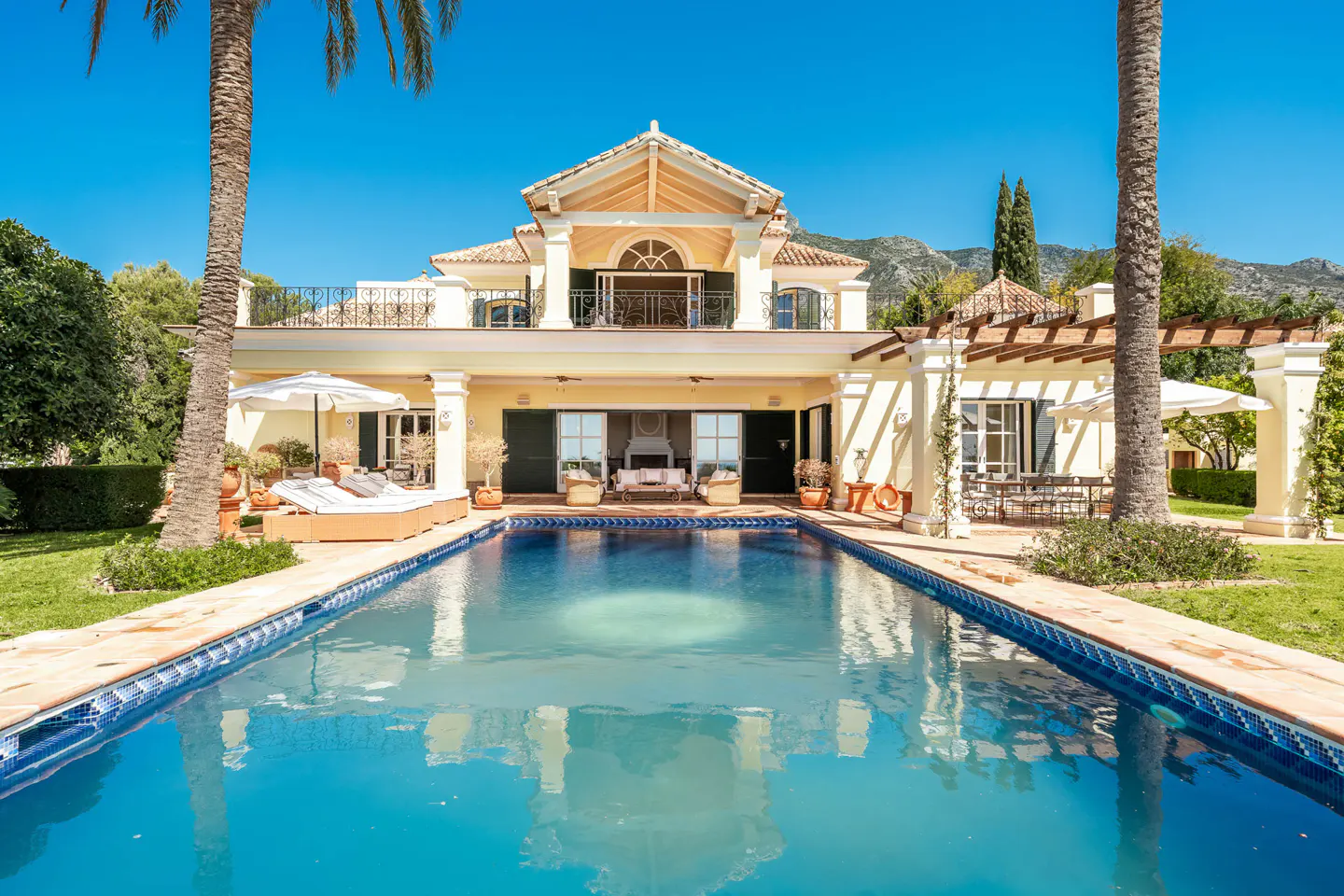 Luxury villa with a blue swimming pool in the foreground, palm trees, and mountains in the background on a sunny day.