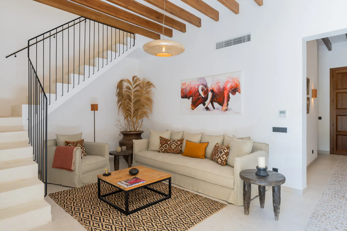 Bright living room with white walls, wooden beams, and staircase. Beige sofa with pillows, patterned rug, and bull painting on the wall.