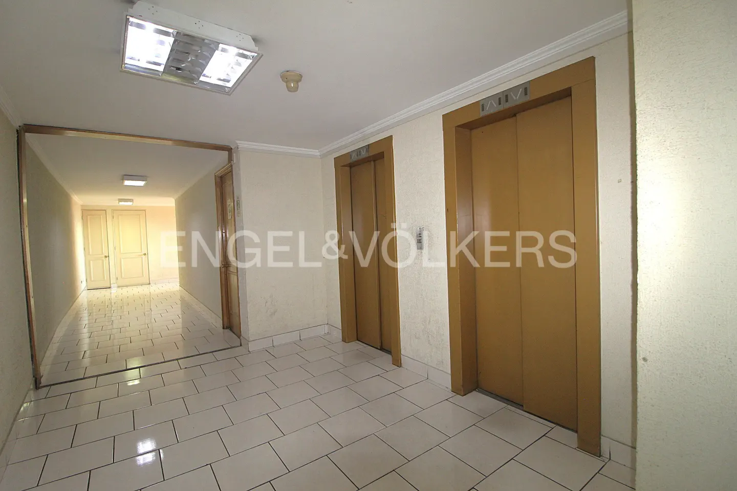 Hallway with two elevators, white tile floor, and a doorway leading to a long corridor with doors.