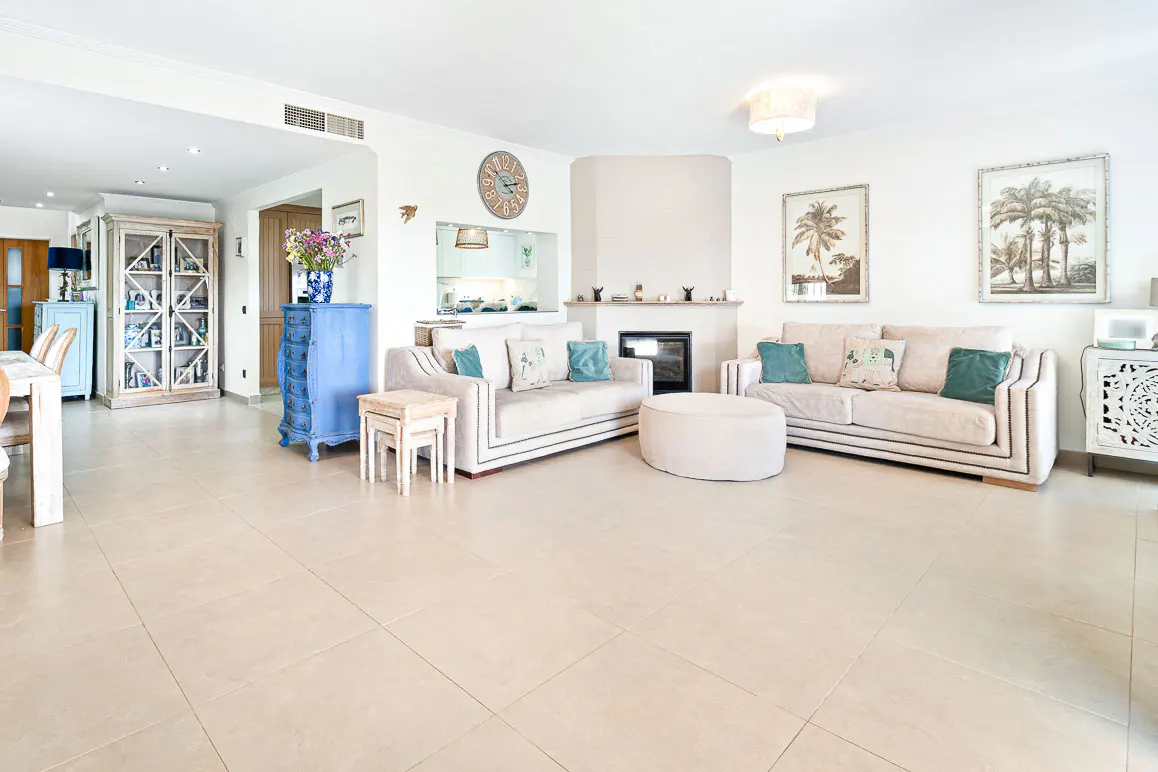 Bright living room with beige sofas, teal pillows, and a fireplace. Palm tree art hangs above. A blue cabinet adds a pop of color.