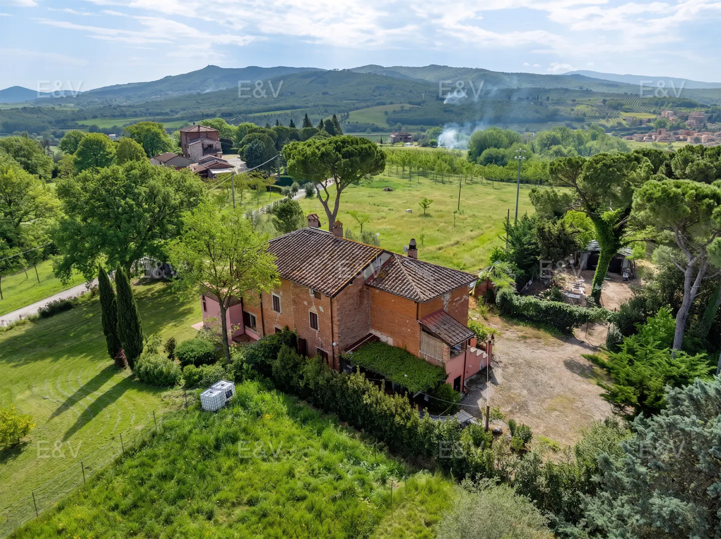 Aerial view of a red brick farmhouse with a tile roof, surrounded by green fields and trees in a rural setting.