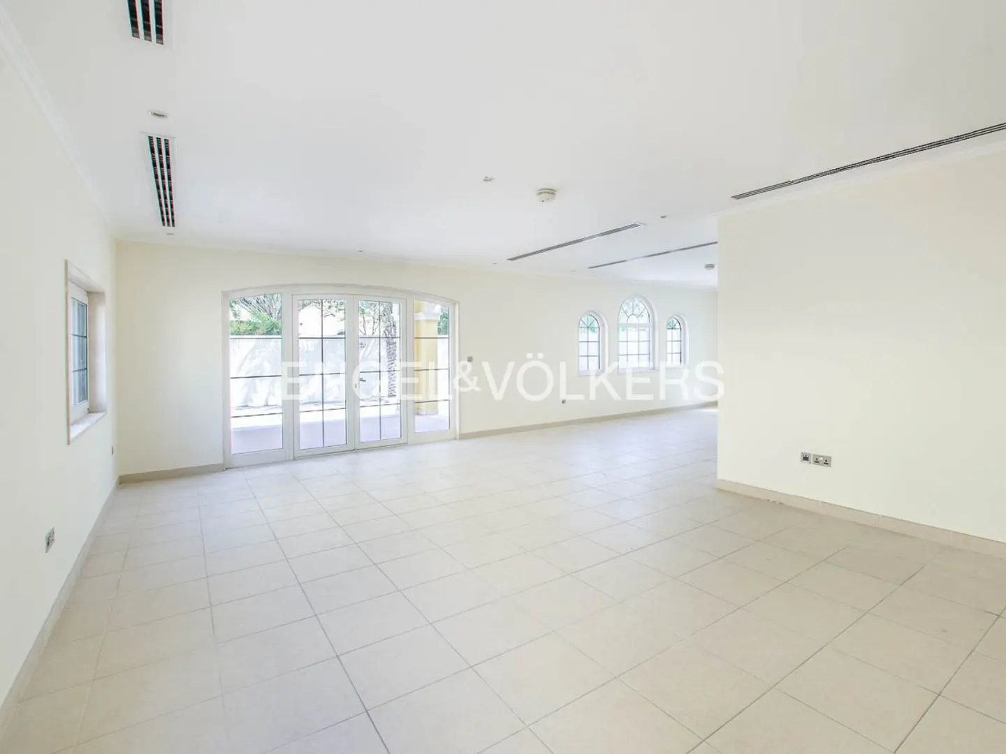Spacious, empty room with white walls, tiled floors, arched windows, and glass doors leading to an outdoor area.