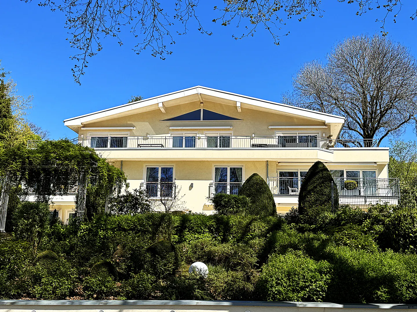Two-story yellow house with white trim, balconies, and green landscaping under a clear blue sky.