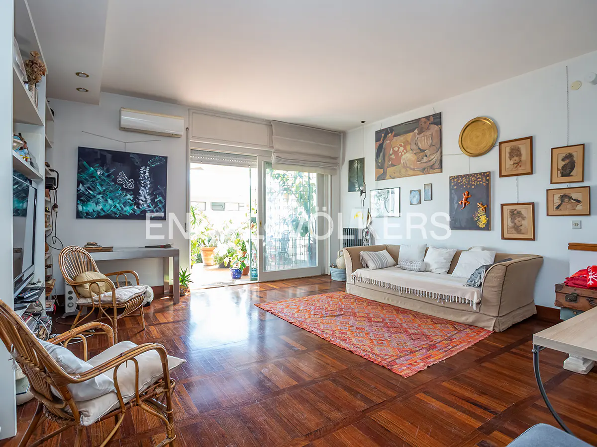Bright living room with hardwood floors, a beige sofa, and a red patterned rug. A balcony with plants is visible through sliding glass doors. Artwork adorns the walls.