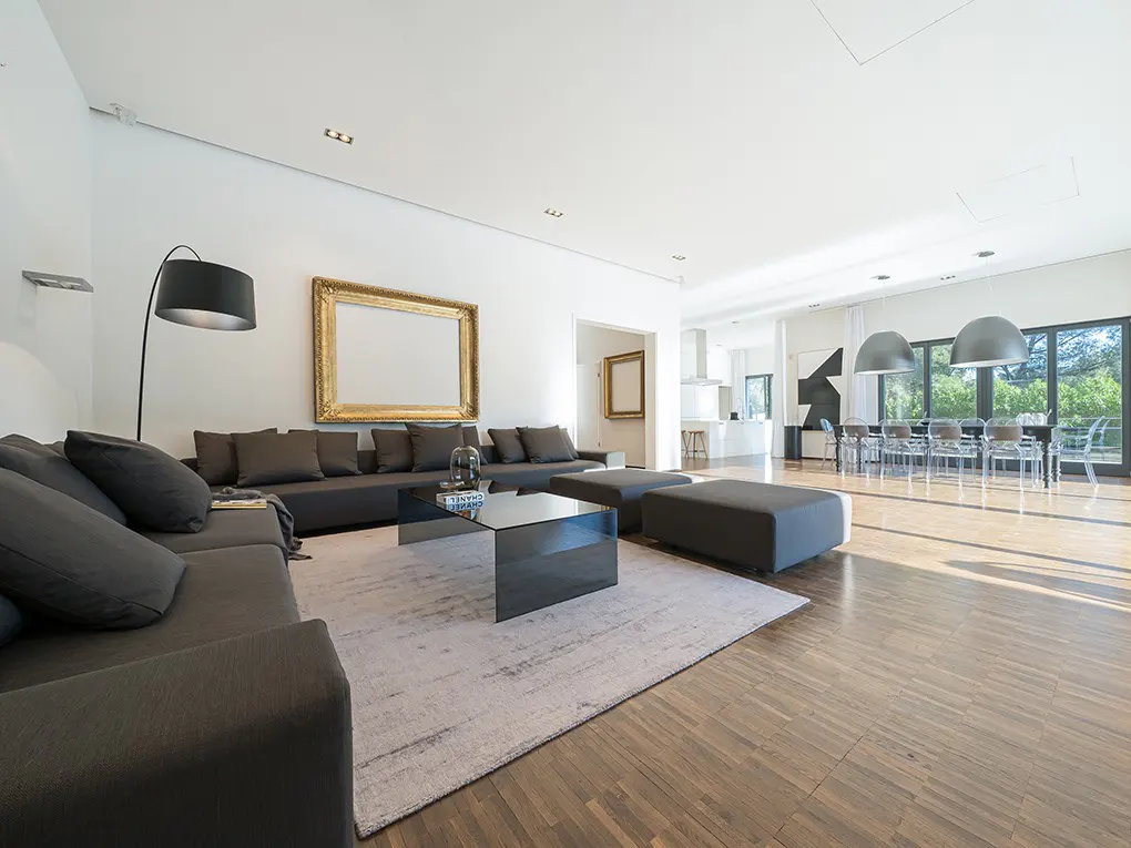 Bright, modern living room with a gray sectional sofa, glass table, and hardwood floors. A large gold frame hangs on the white wall.
