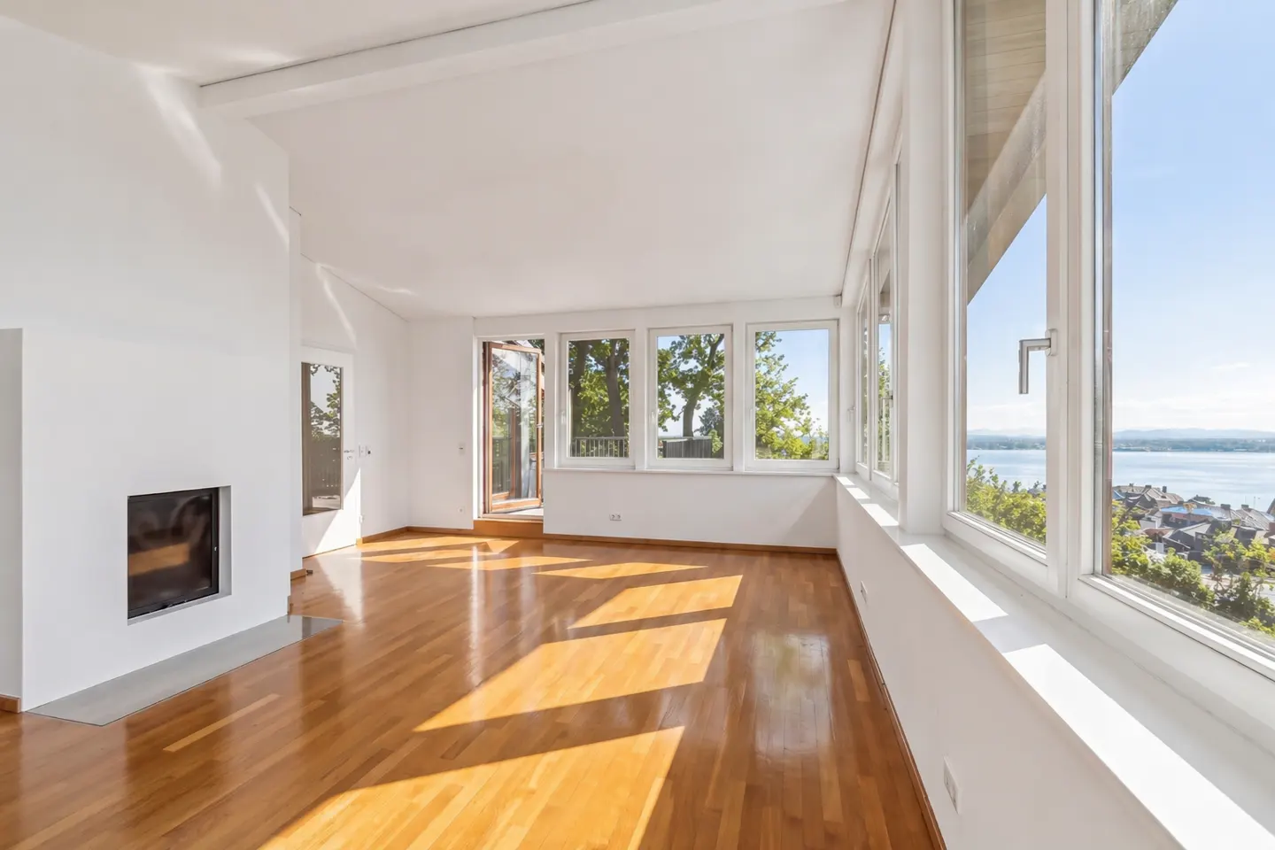 Bright, empty living room with hardwood floors, white walls, fireplace, and large windows with a view of trees and water.