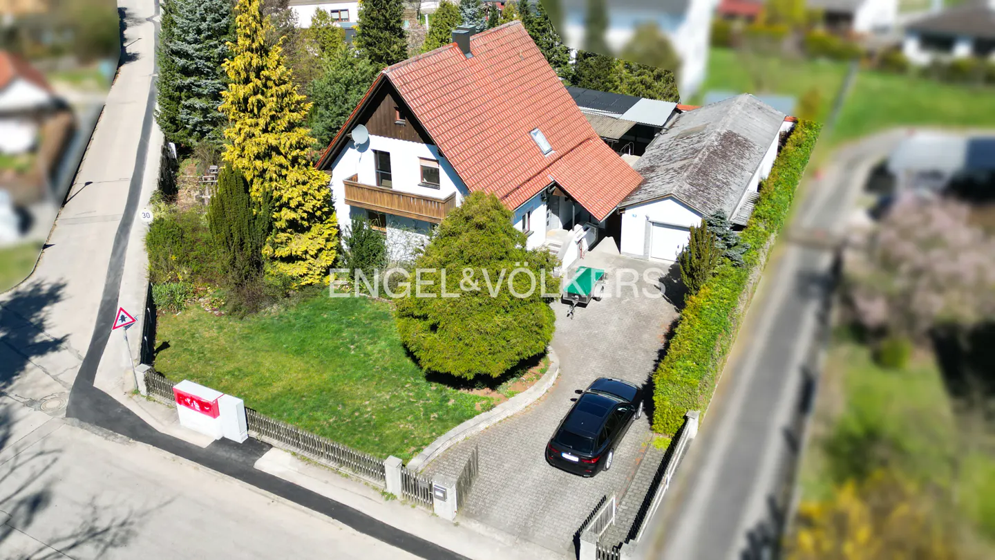 Aerial view of a white house with a red tile roof, green lawn, and a black car parked in the driveway. The Engel & Völkers logo is visible.