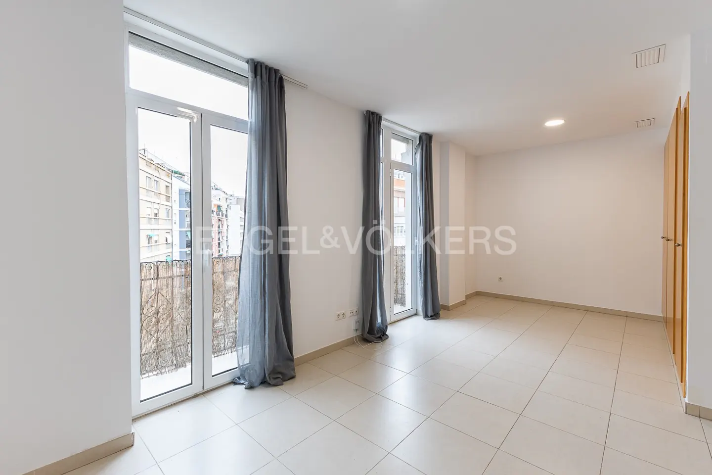 Bright, empty room with white walls, tiled floor, and two sets of balcony doors with gray curtains.