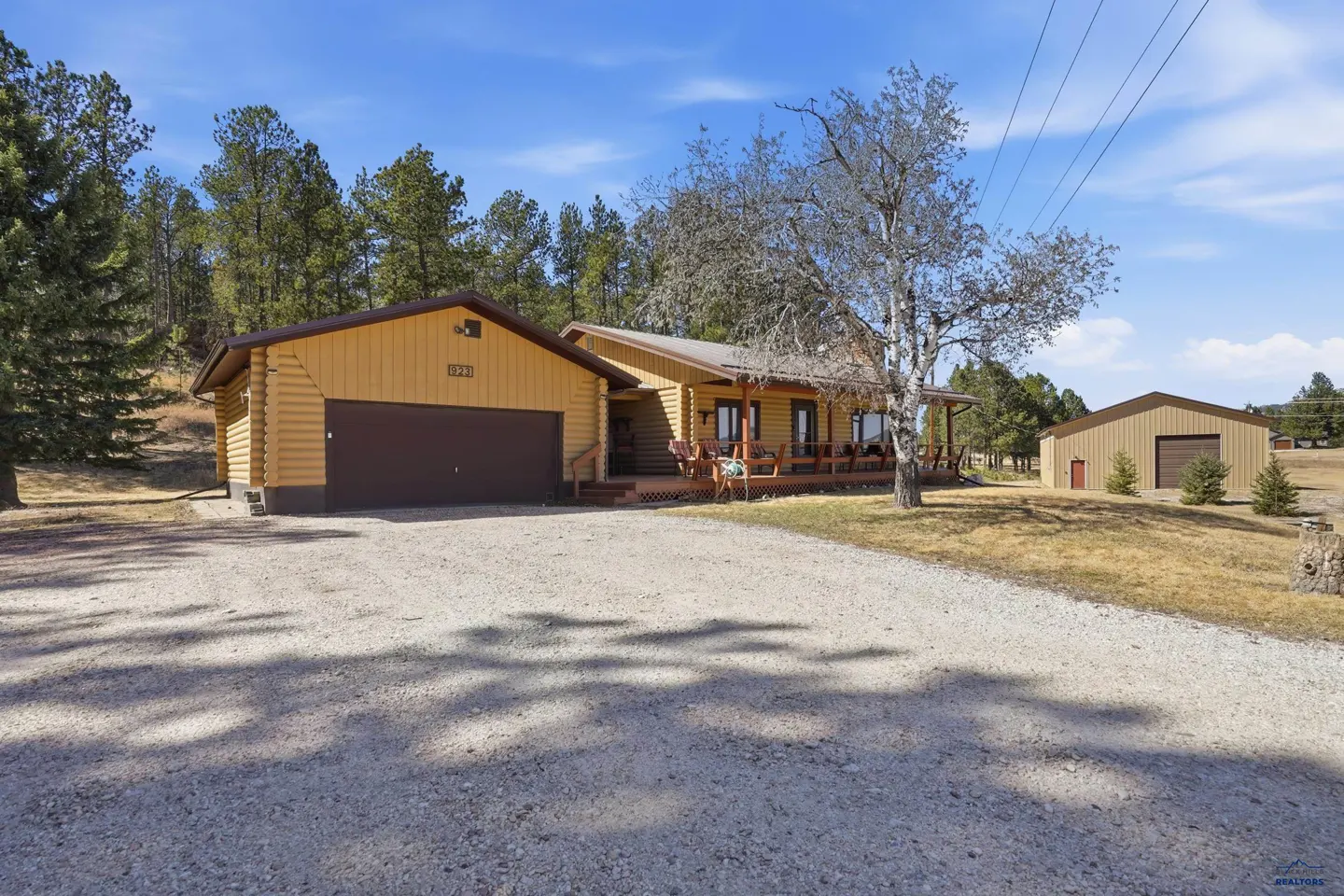 A tan log cabin home with a brown garage door and a gravel driveway. A large tree stands to the right of the house.