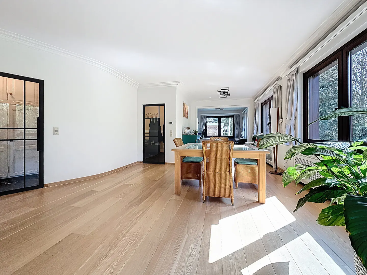 Bright, airy dining room with light wood floors, white walls, and a wooden table with wicker chairs. Black framed doors and large windows add contrast.
