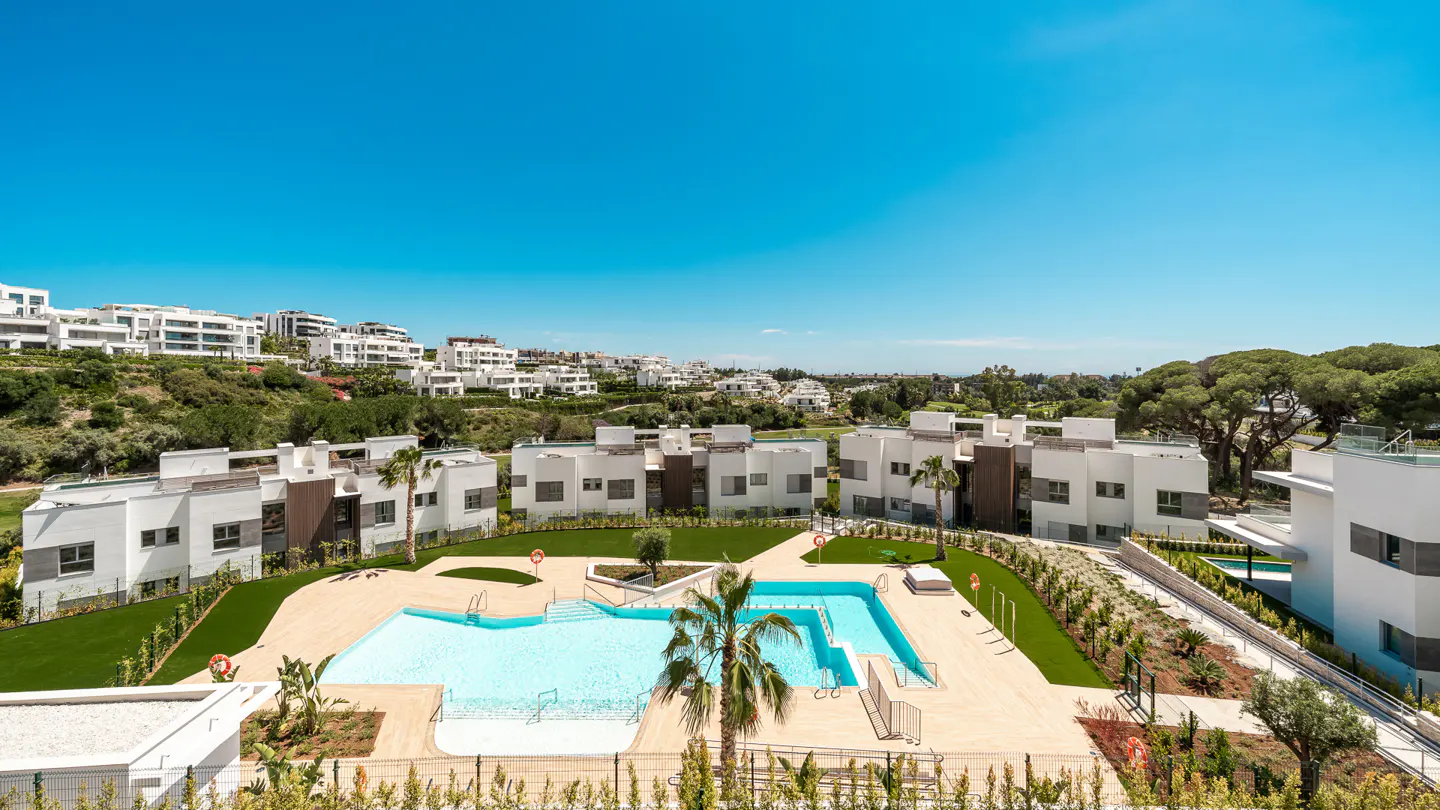 Aerial view of a modern white apartment complex with a turquoise pool and palm trees under a clear blue sky.