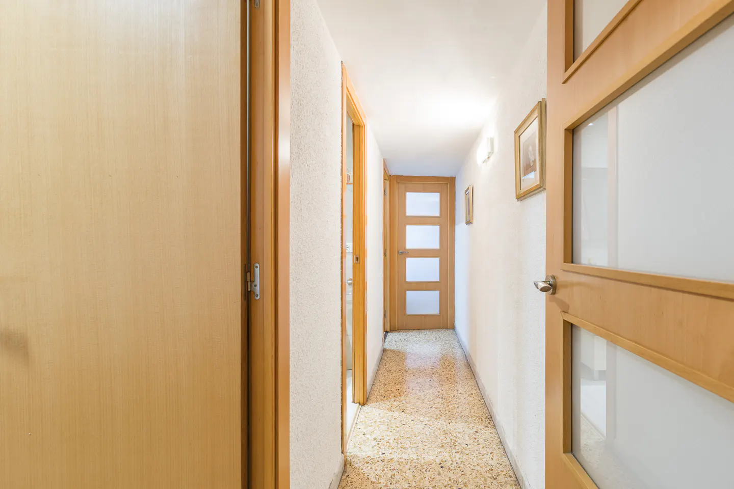 Hallway with light wood doors, white walls, and a speckled floor. A door with glass panels is partially open on the right.