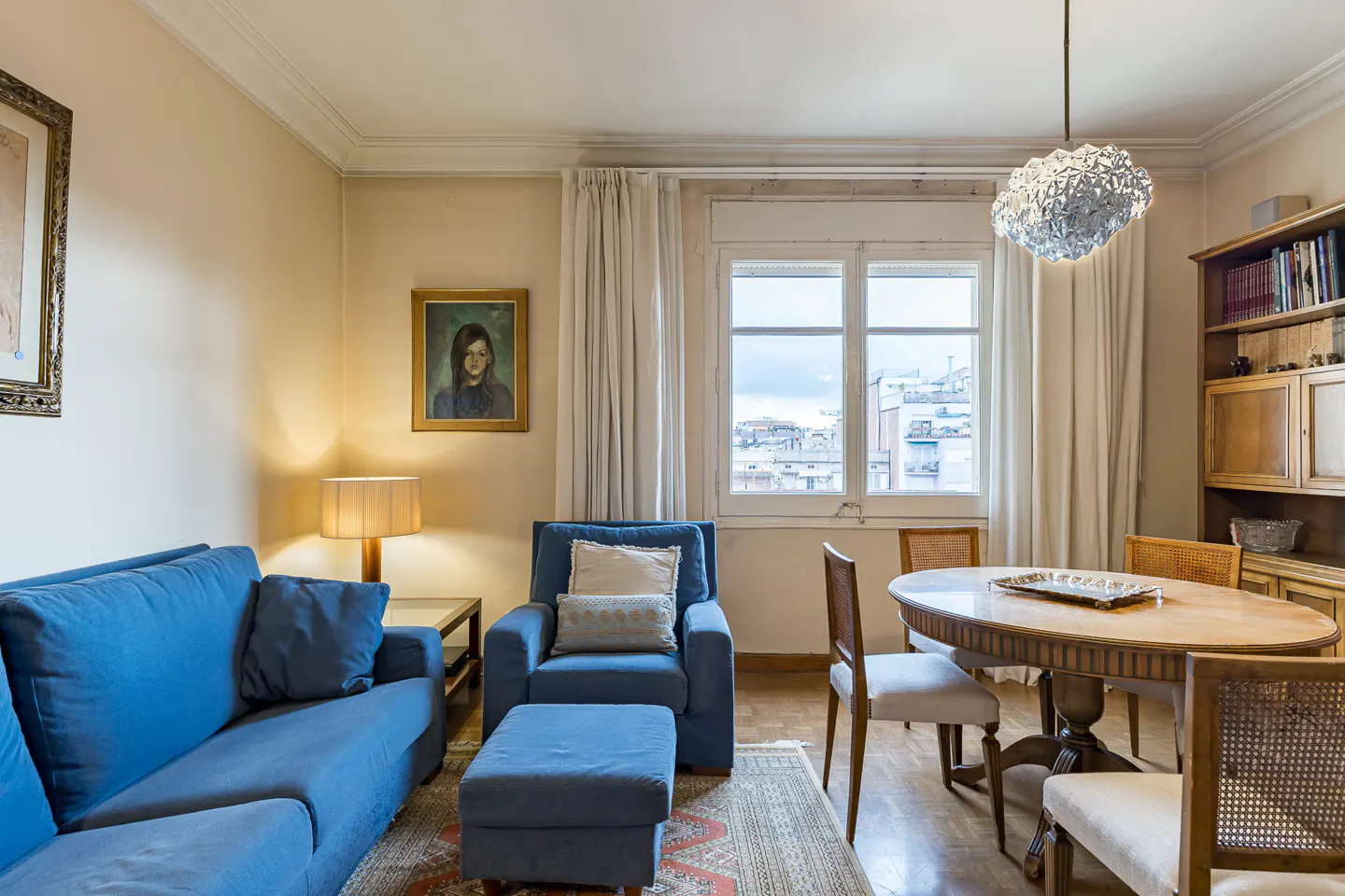 Living room with blue sofa, armchair, ottoman, and wooden dining table with chairs under a modern crystal pendant light.