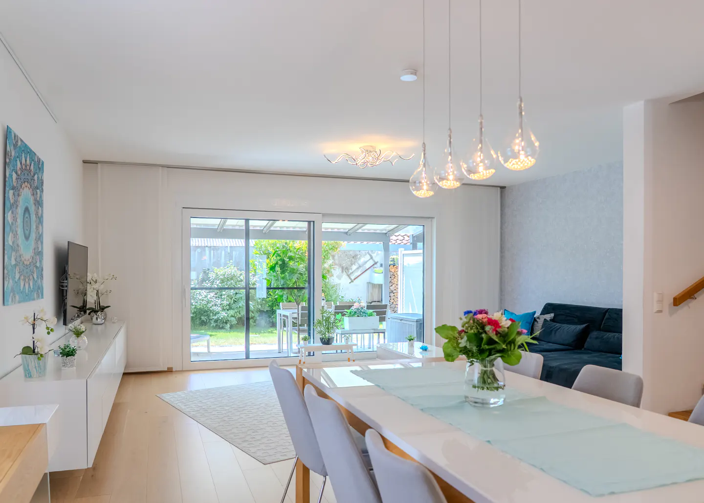 Bright dining room with a white table, light blue runner, and pendant lights, opening to a patio with a garden view.
