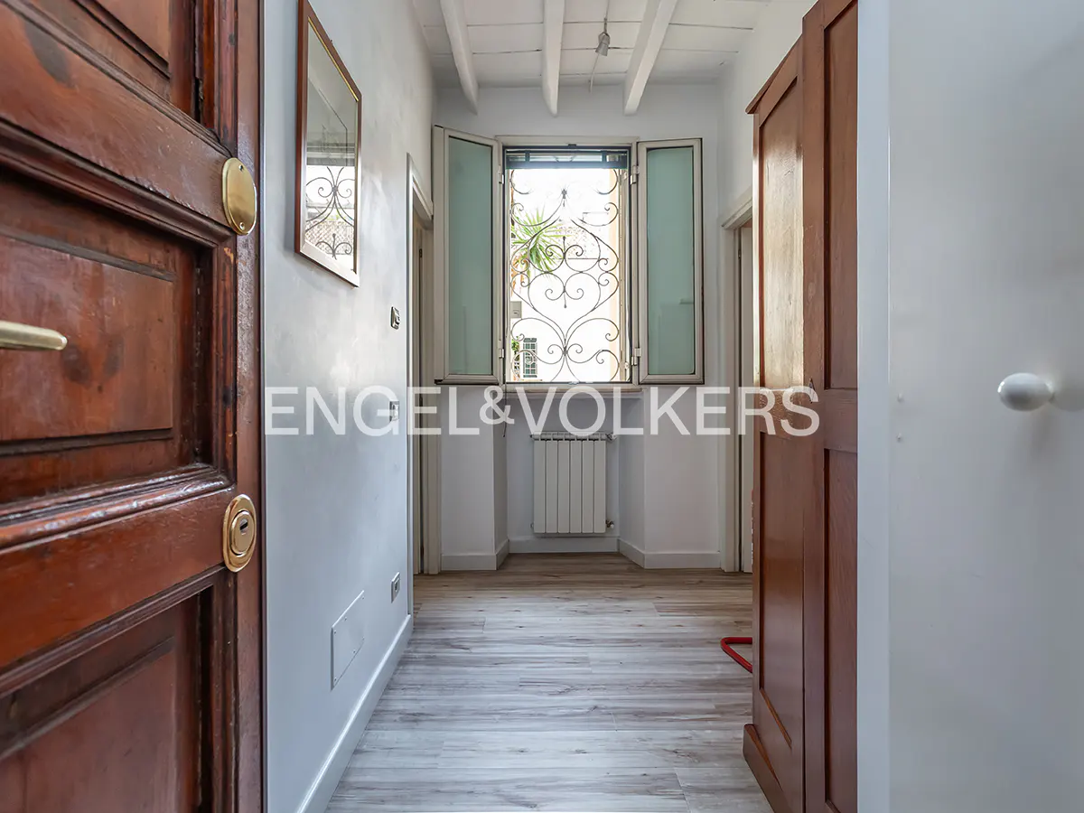 A bright hallway with white walls, a wooden floor, and a window with decorative ironwork. A dark wood door is on the left.