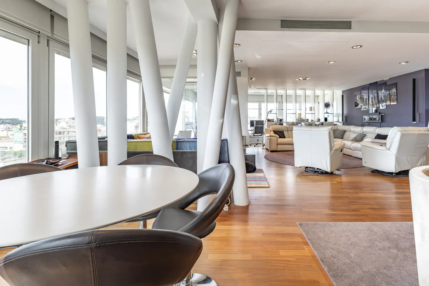 Bright, open-concept living space with hardwood floors. A white table and brown chairs sit near large windows and white support columns. Sofas and decor are in the background.