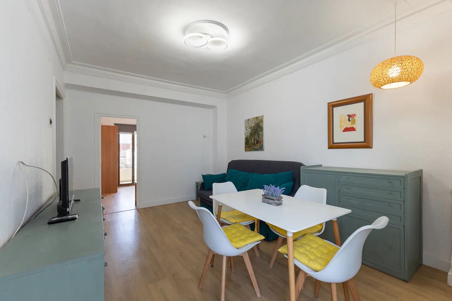 Bright living room with white walls, wood floors, and blue-green accents. A white table with yellow cushions sits near a gray sofa with teal pillows.