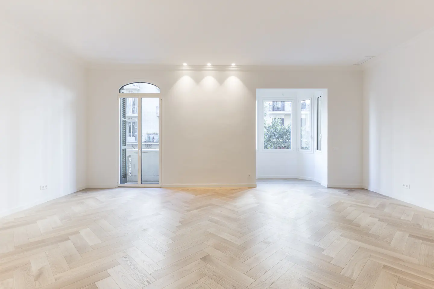 Bright, empty room with white walls, herringbone wood floor, arched balcony door, and windows with city views.