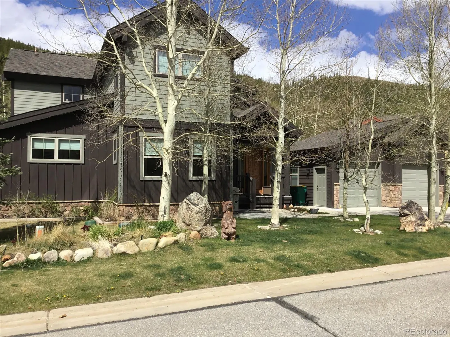 Two-story house with brown siding and gray trim, surrounded by birch trees and a green lawn. A mountain is visible in the background.