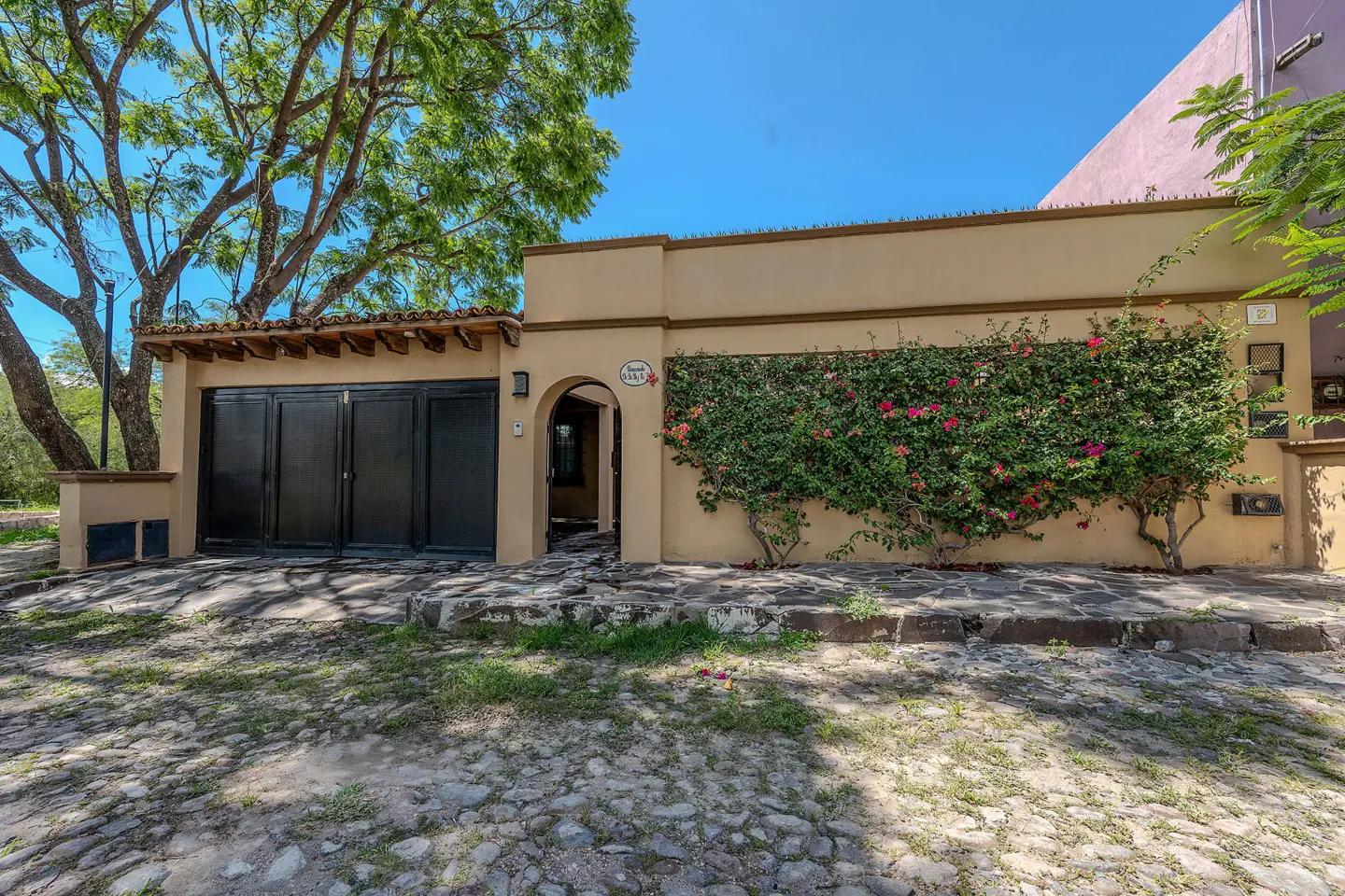 Tan house with black garage door and arched doorway, covered in flowering vines. Cobblestone street in front.