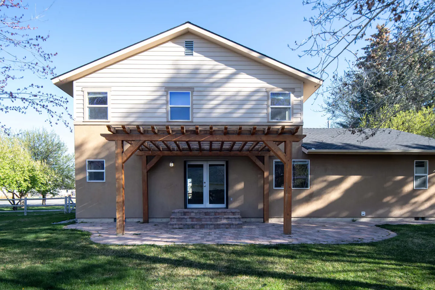Back of a two-story house with a wooden pergola over a patio. The house is beige with white trim and windows. Green lawn and trees surround the house.