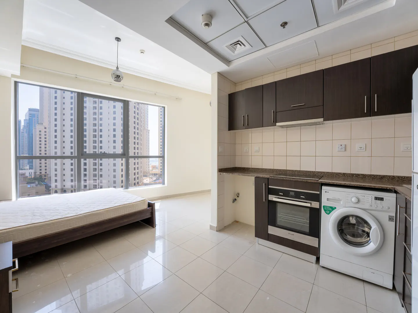 Studio apartment with a bed, kitchen cabinets, oven, and washing machine. A large window shows a city view.