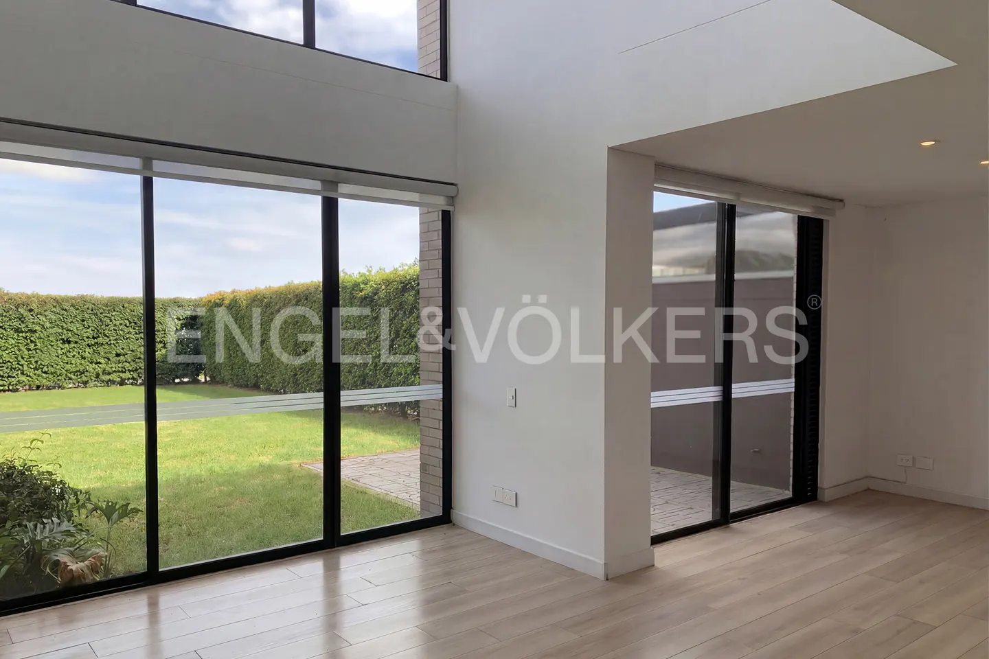Bright, empty room with light wood floors, large windows, and white walls. Green lawn and hedges visible outside. Engel & Völkers logo on window.