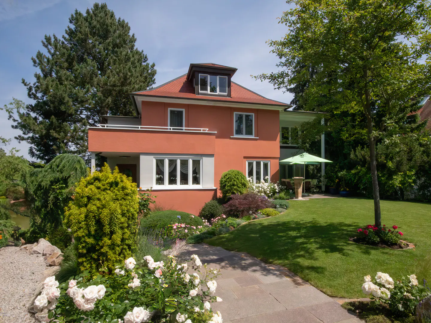 Two-story orange house with a red roof, white windows, and a stone path leading to the front door. Lush green lawn and trees surround the house.