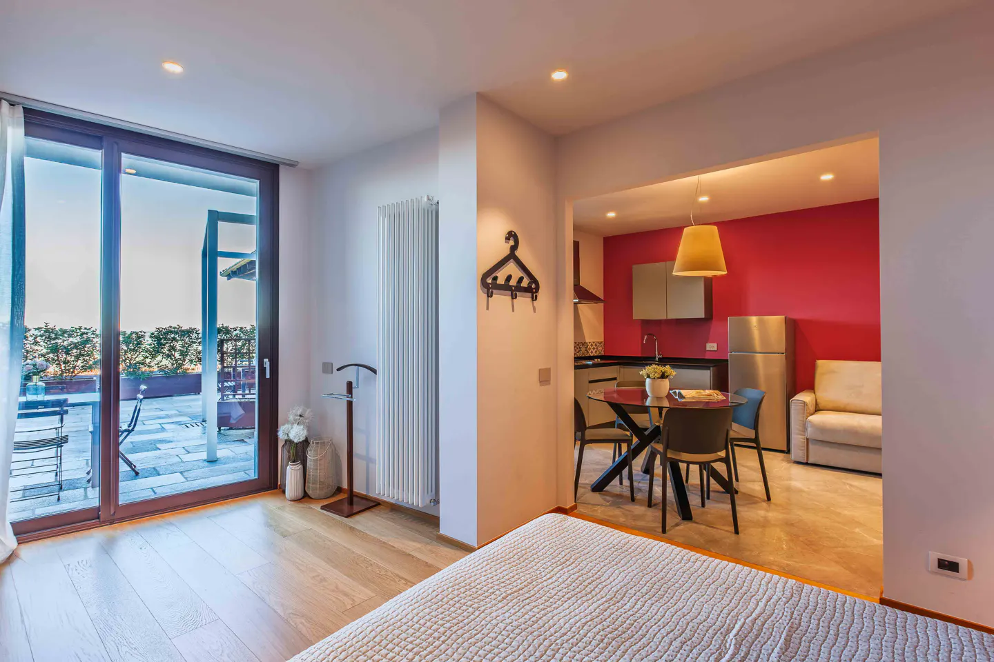 Bright studio apartment with a balcony. A kitchen with red wall, table, and sofa is visible. The balcony has outdoor furniture.