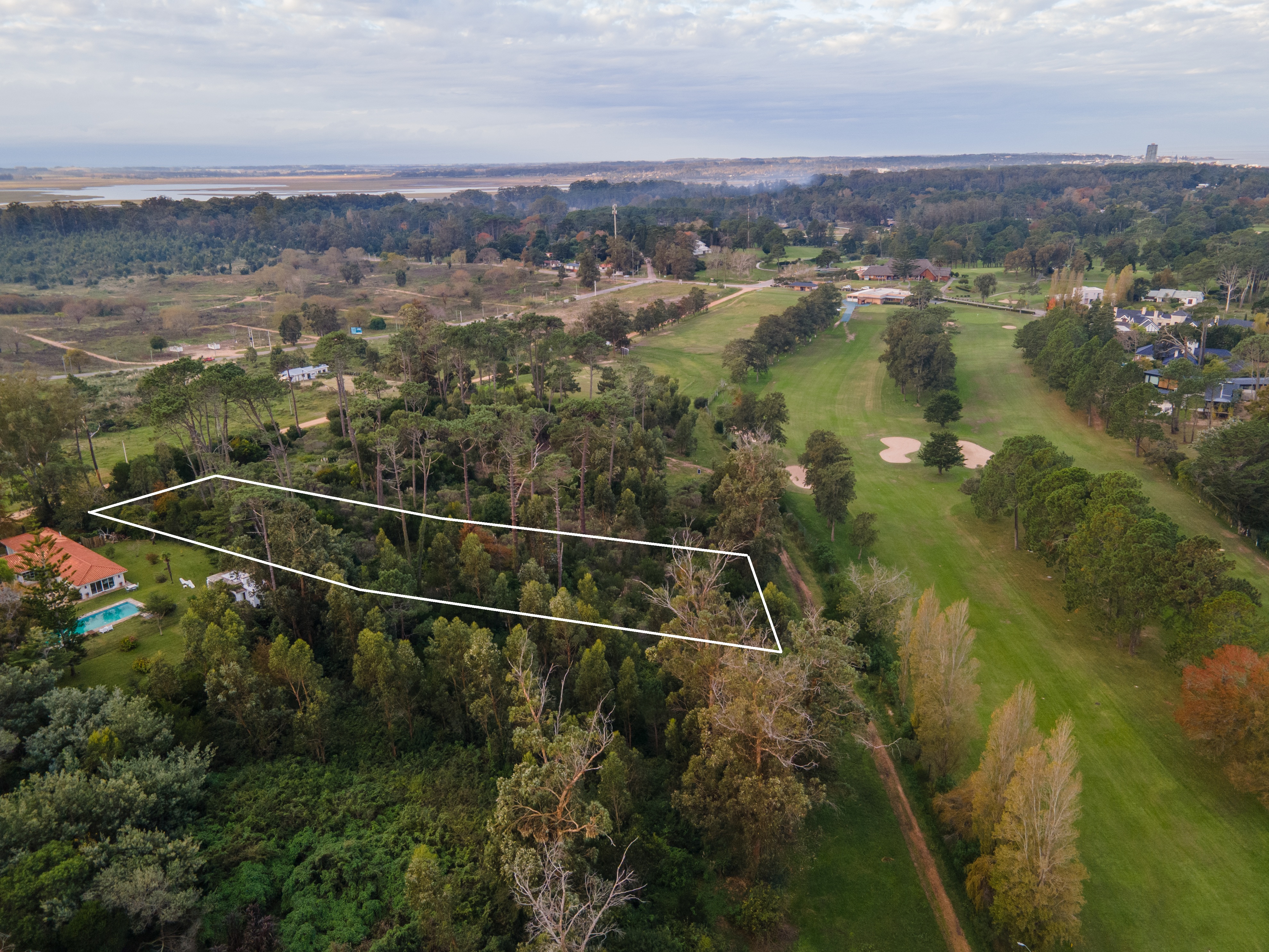 Aerial view of a wooded lot outlined in white, next to a golf course and a red-roofed house with a blue pool.