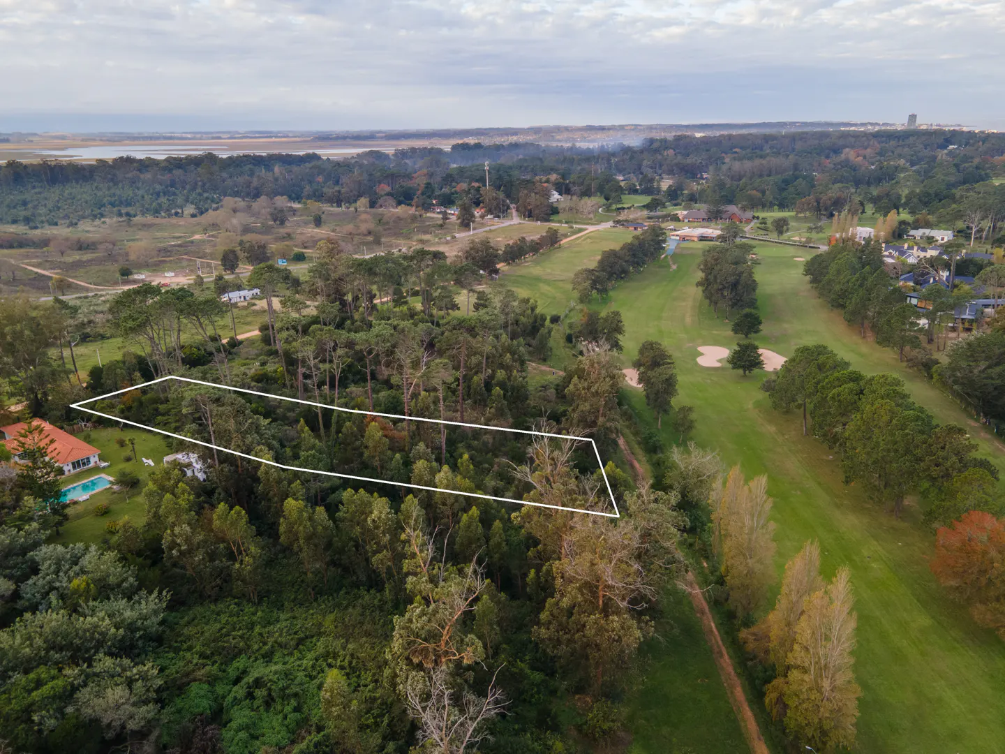 Aerial view of a wooded lot outlined in white, next to a golf course and a red-roofed house with a blue pool.
