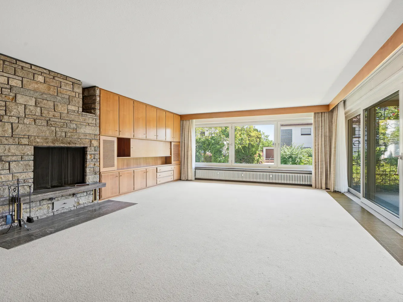 Spacious living room with a stone fireplace, light wood built-in cabinets, white carpet, and large windows with garden views.