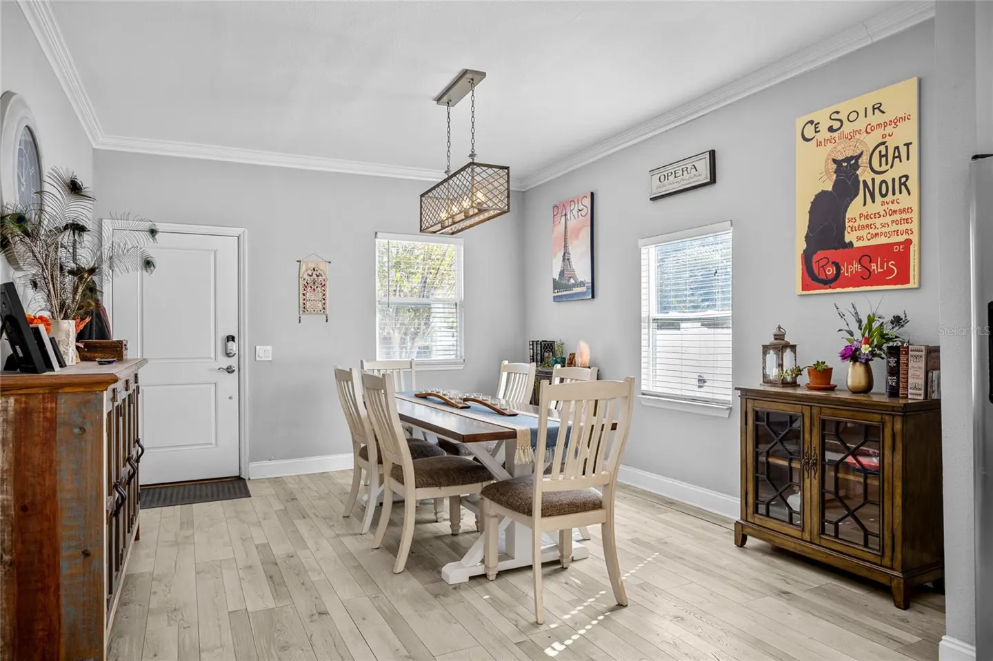 Dining room with a wooden table, six chairs, and a metal chandelier. Artwork hangs on the gray walls. A wooden cabinet sits near a window.