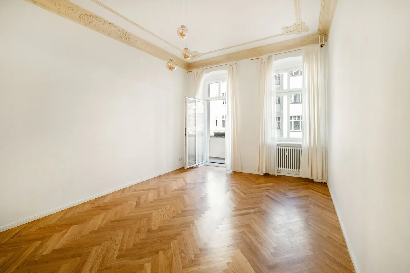 Bright, empty room with herringbone wood floors, white walls, and tall windows with sheer curtains. A balcony door is open.